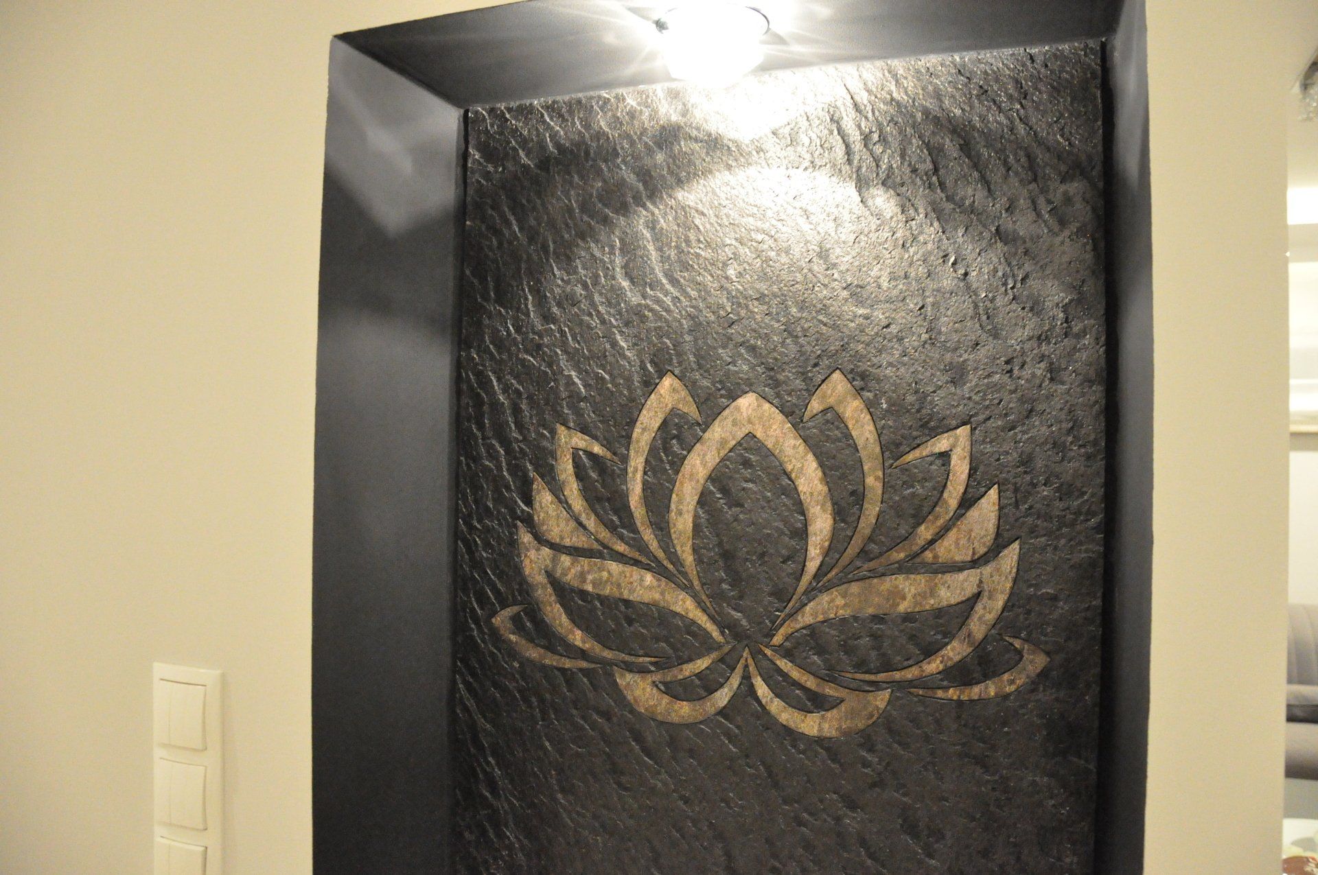 Niche wall with a gold lotus flower design on a dark textured background, lit by a spotlight.