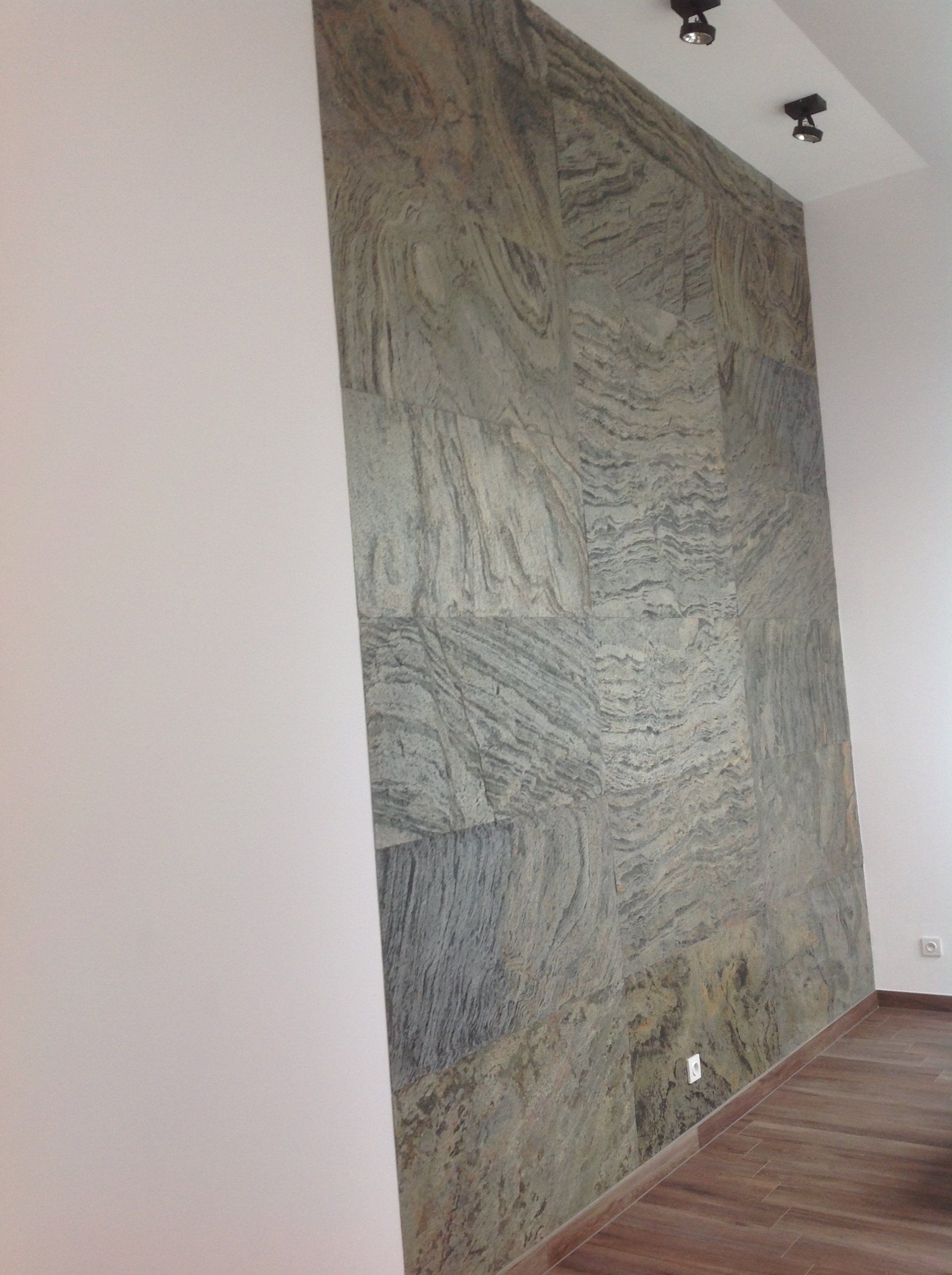 Stone-tiled accent wall in a room with white walls and a brown floor. Spotlights are on the ceiling.