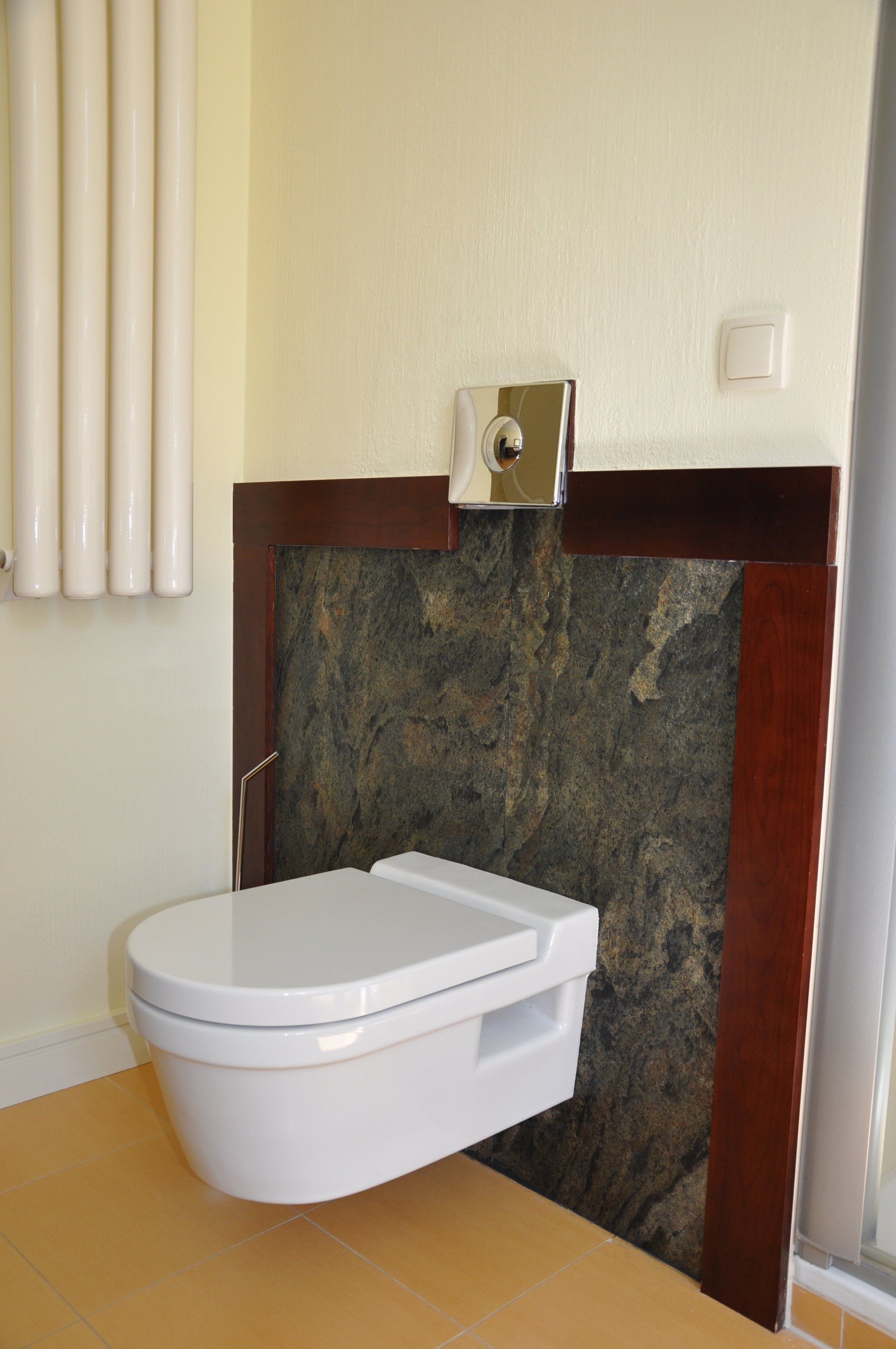 Wall-mounted white toilet against a dark stone backsplash framed in wood. Silver flush plate above. Light yellow walls.