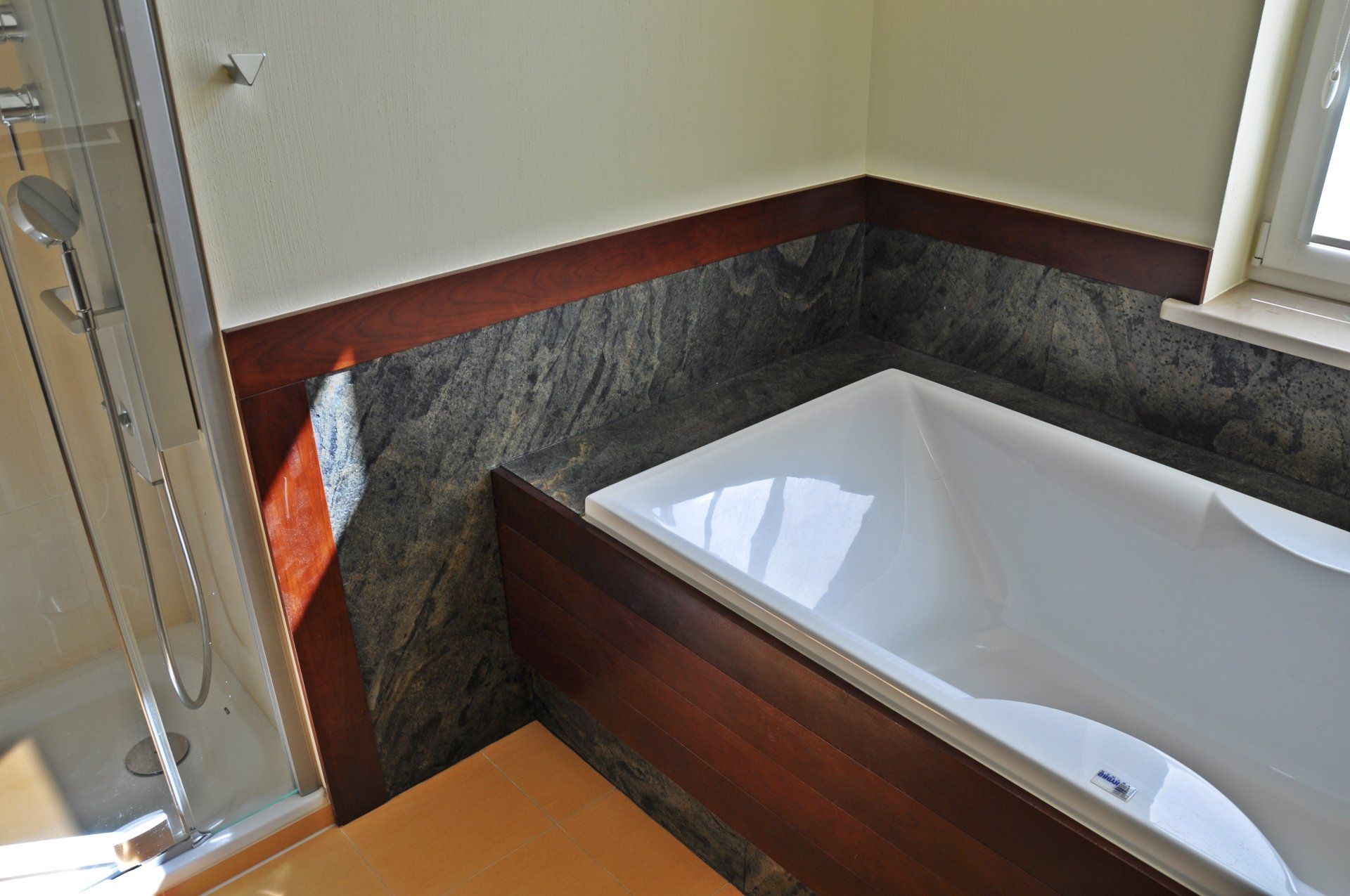 Bathroom with white bathtub, granite wall, and wood trim. A shower is on the left.