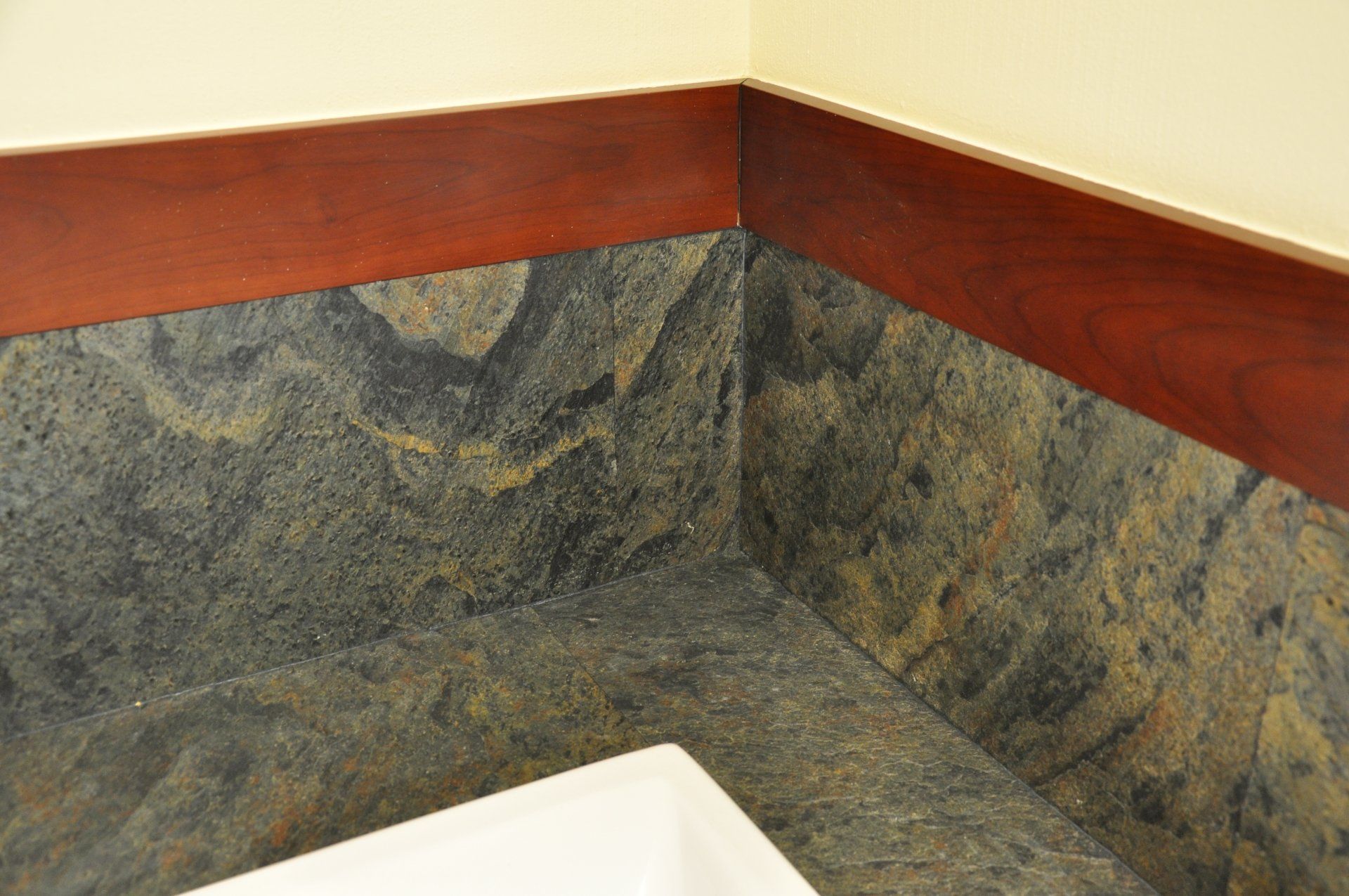 Corner of a wall with dark slate-like tiles and a stained wood trim, near a white surface.