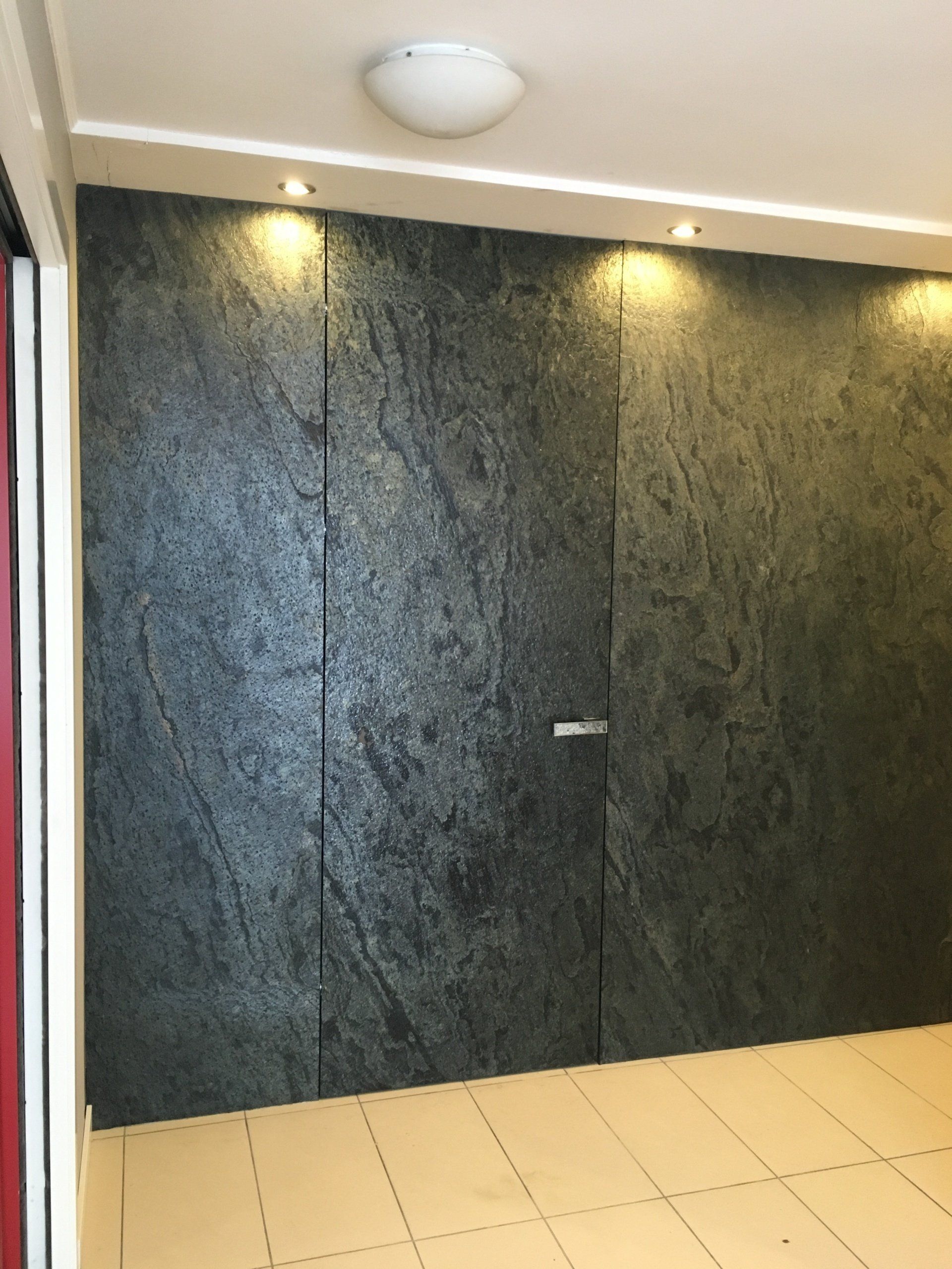 Dark stone-like wall with a hidden door, illuminated by spotlights and a ceiling light.