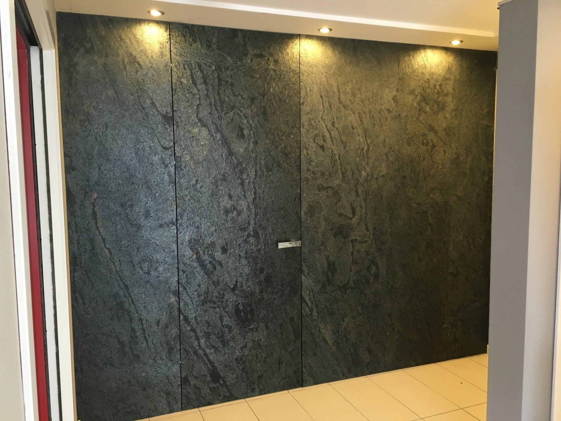 Dark gray textured stone wall with hidden door. Warm lighting from above.
