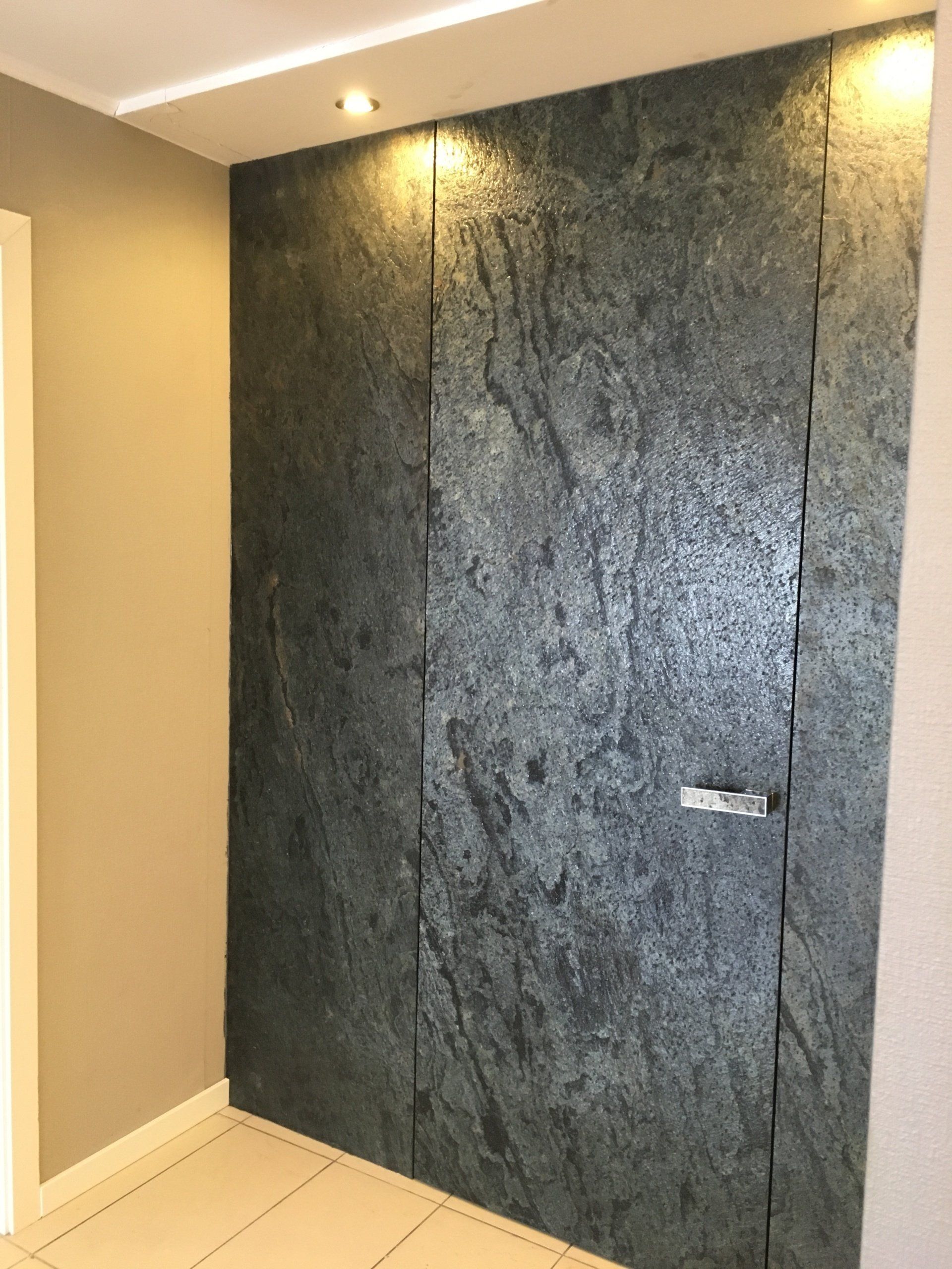 A dark, stone-paneled door in a hallway. It is a flush-mount design with a silver handle.
