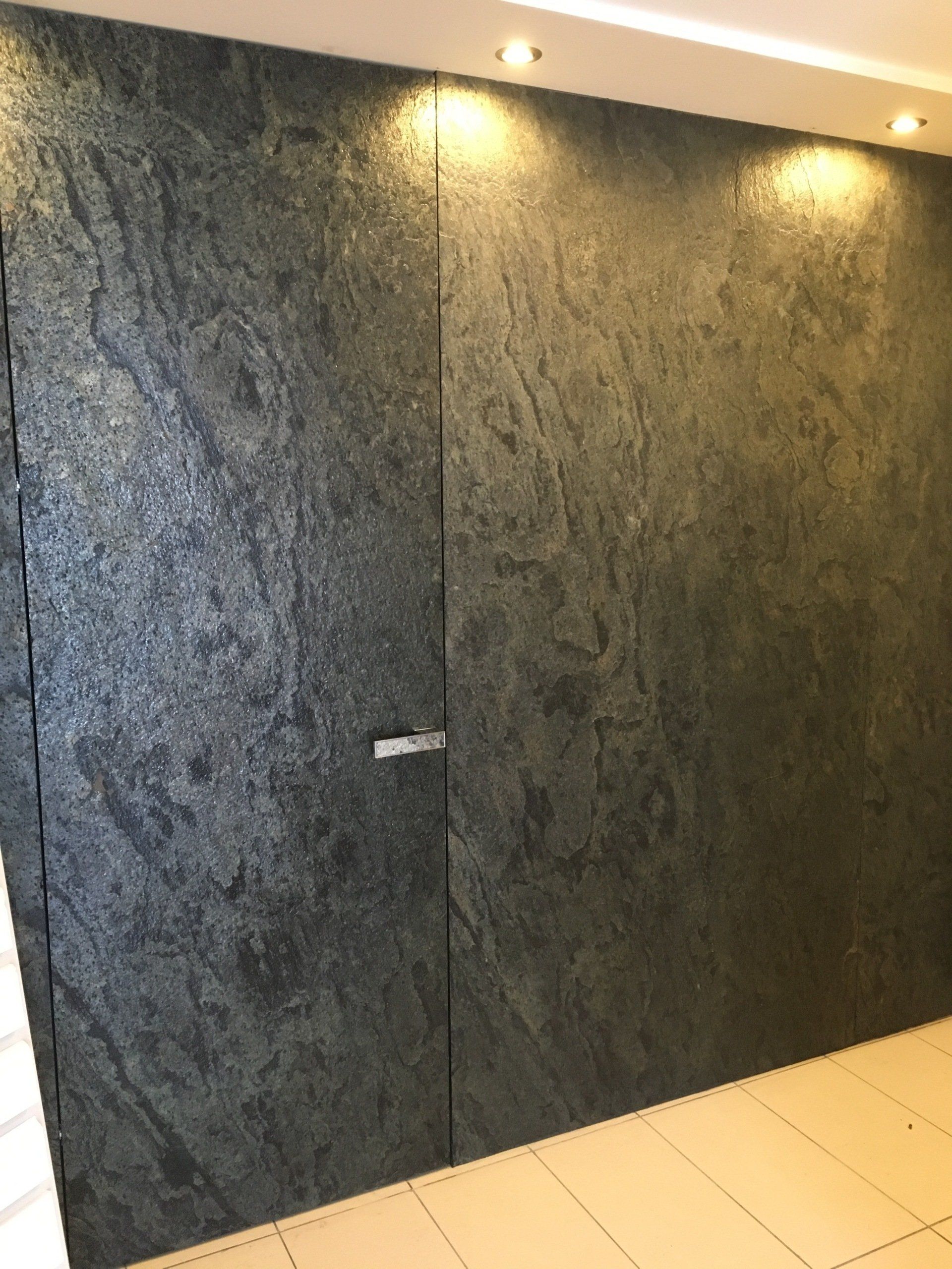 Dark gray stone wall with two panels, one being a hidden door, lit by recessed lights.