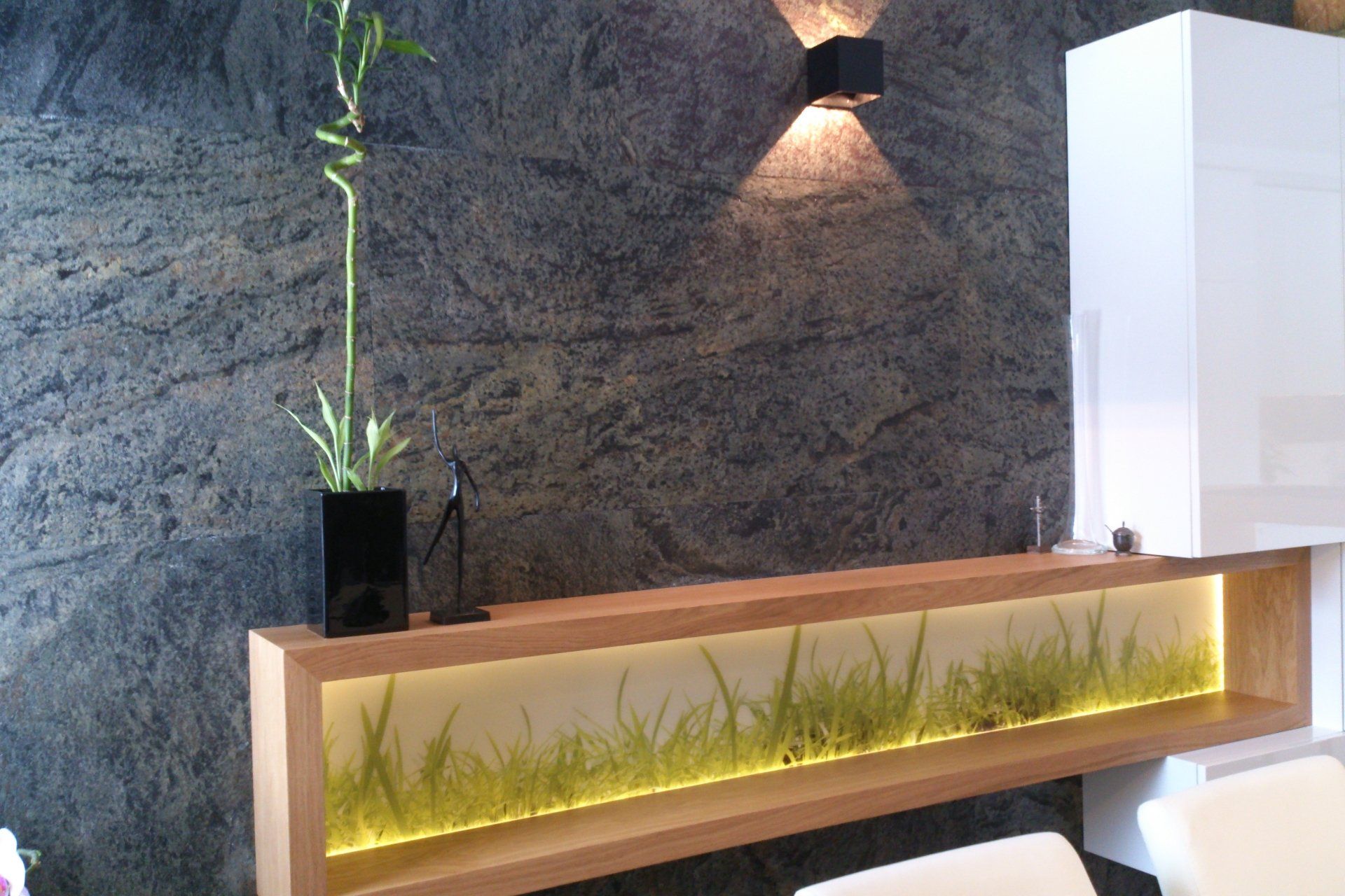 Wooden shelf with illuminated green grass, black vase with bamboo, against a textured gray wall.