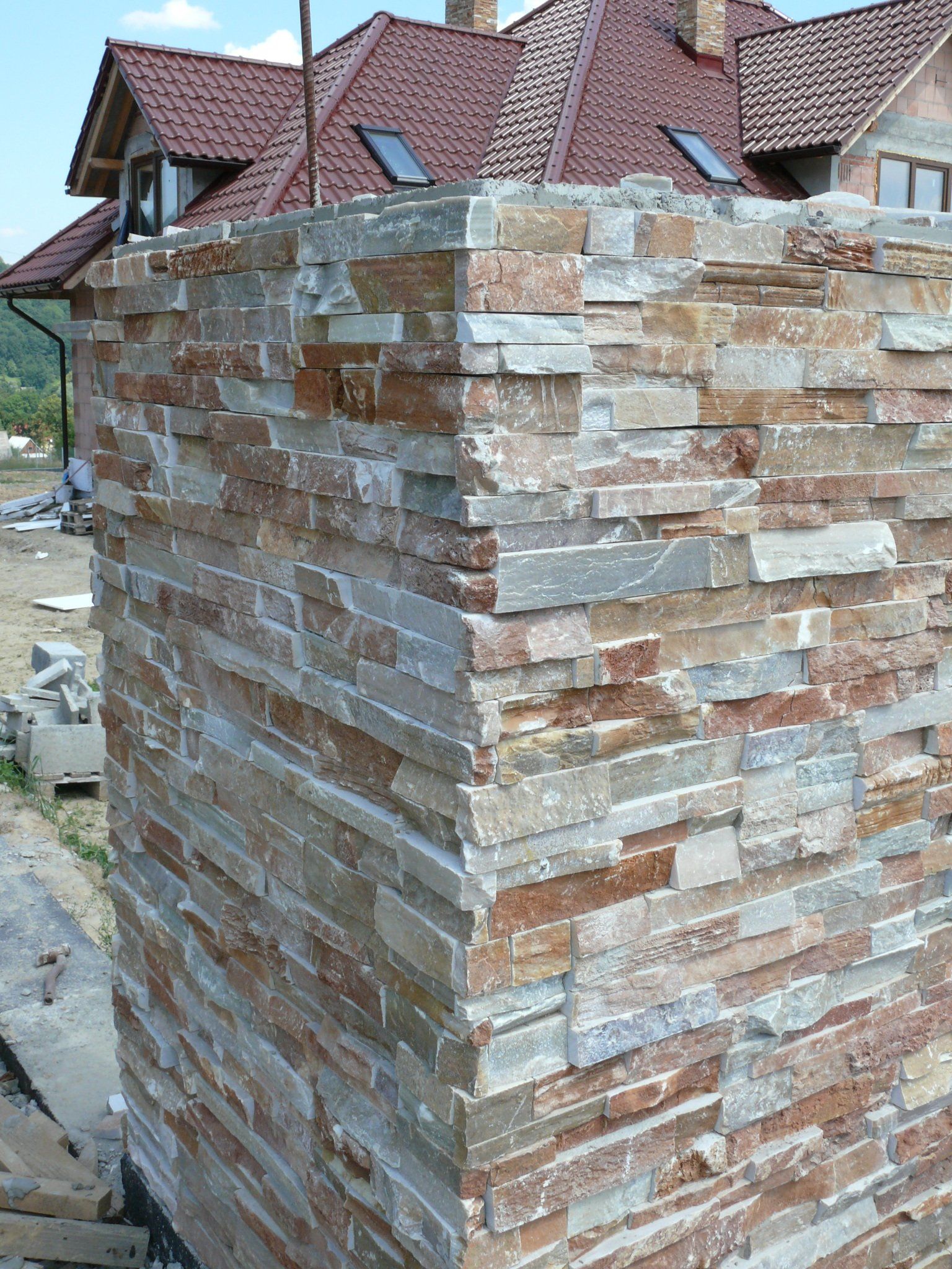 Stone veneer on a corner wall, featuring varied reddish-brown and gray tones, with a house and roof in the background.