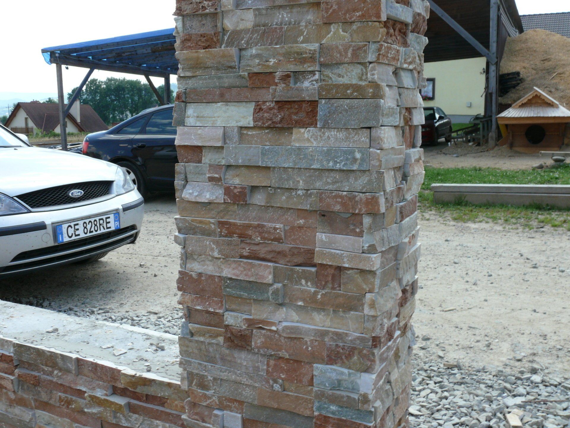 Stone-clad pillar in a driveway with cars and a building in the background.