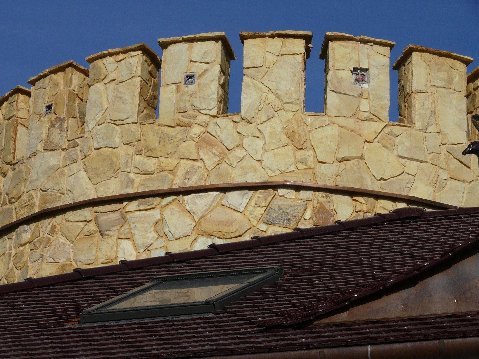 Stone castle wall with battlements above a brown tiled roof, against a blue sky.