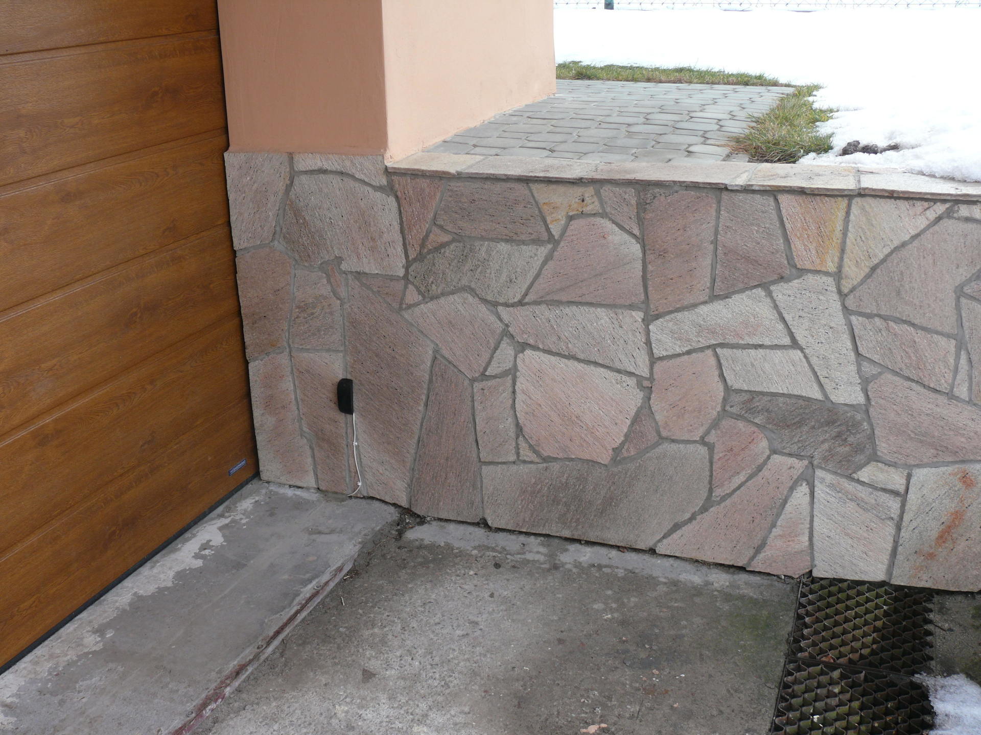 Stone-covered wall section next to a wooden garage door; concrete porch with a doormat; snowy ground.