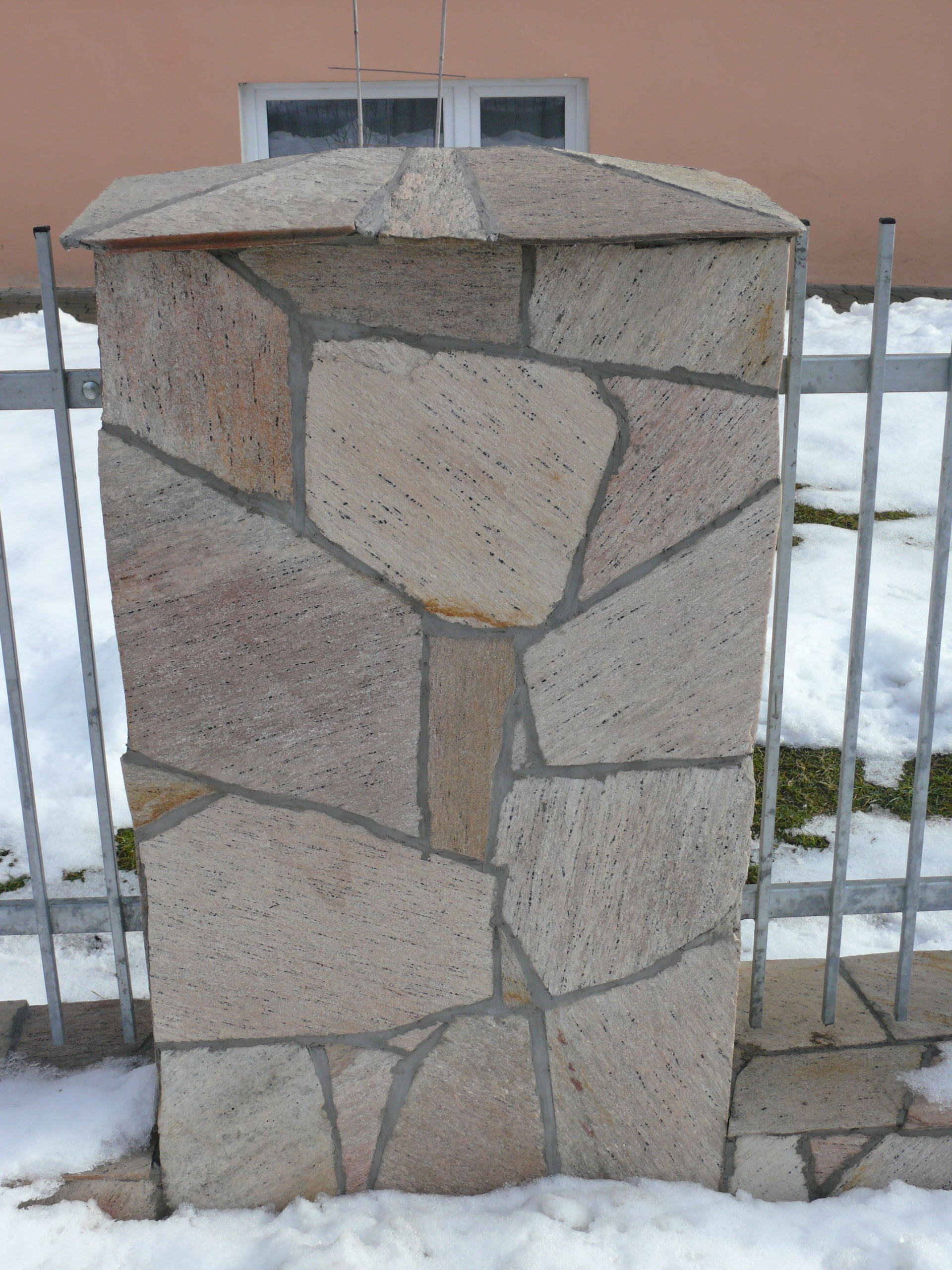 Stone pillar with a decorative cap and sections, supporting a metal fence with snow on the ground.