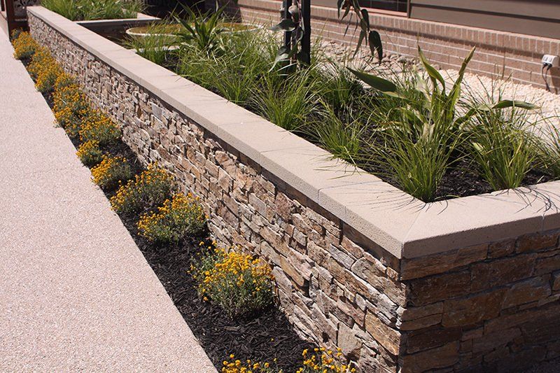 Stone wall planter with yellow flowers and green plants, next to a concrete sidewalk.