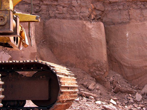 Excavator tracks and arm next to a red-brown rock face with carved rectangular shapes.