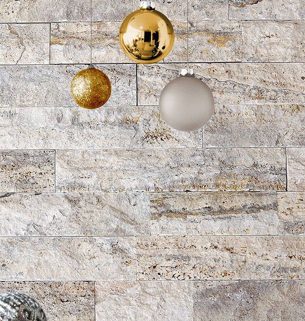 Three Christmas ornaments hanging against a textured stone wall: gold, sparkly gold, and silver.