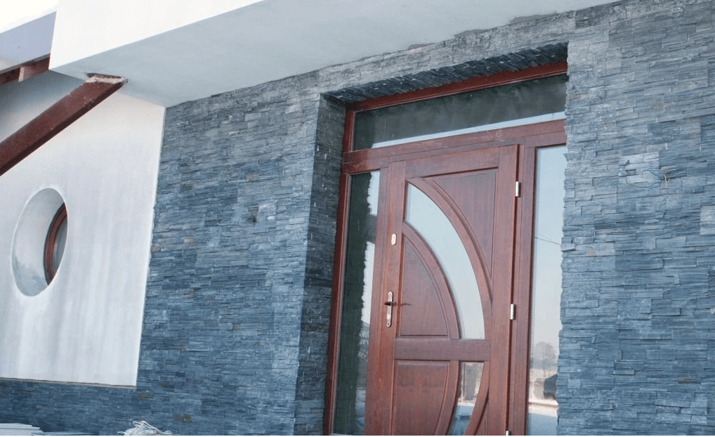 Exterior of a building with stone facade, brown wooden door with glass panels, white wall with circular window.