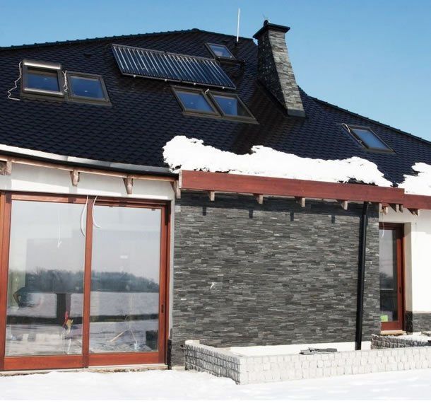 House with black roof, solar panels, snow, and stone wall; winter setting.
