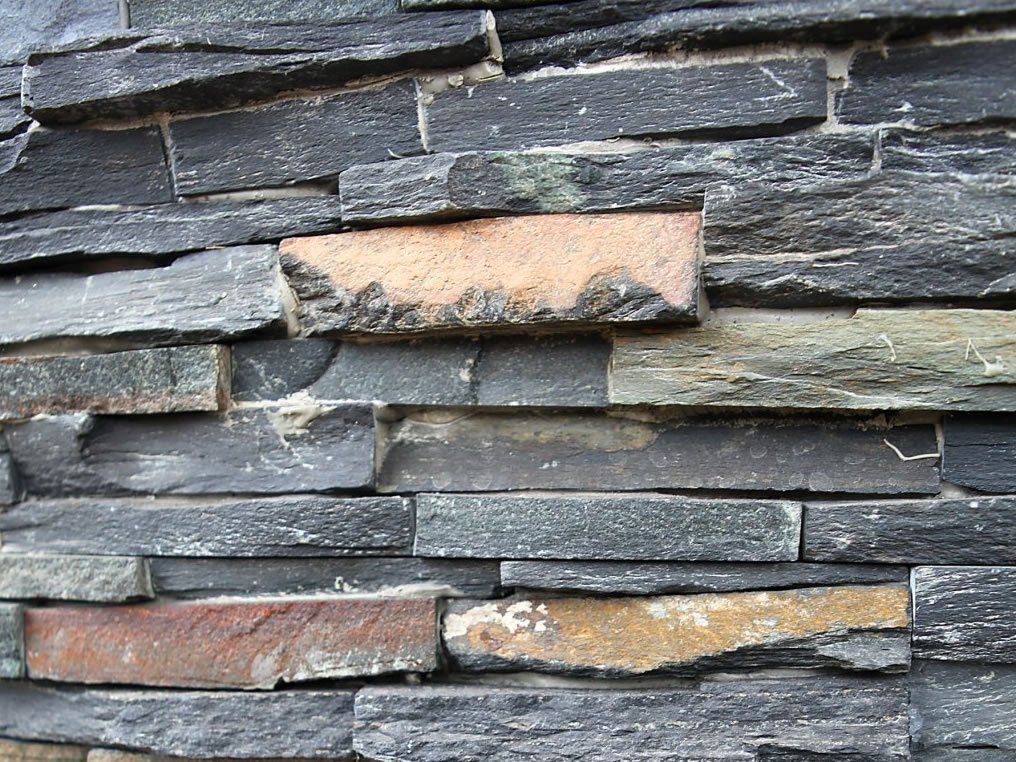 Close-up of a stone wall. Mostly dark gray and black slate with a few tan and reddish-brown stones.