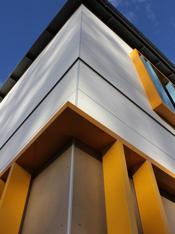 Corner of a modern building with silver panels and bright yellow architectural accents, against a blue sky.