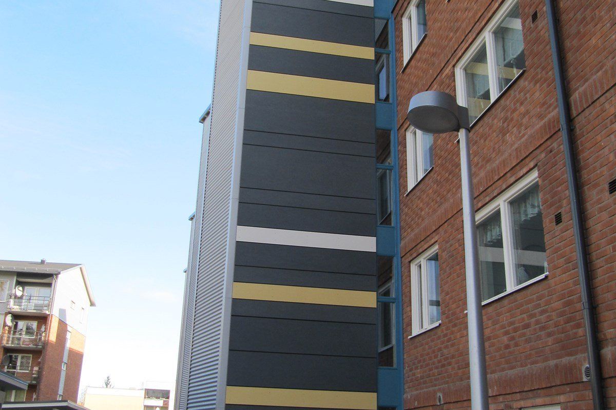 Tall building with a colorful facade: dark grey, yellow, and white stripes. A red brick building is on the right.