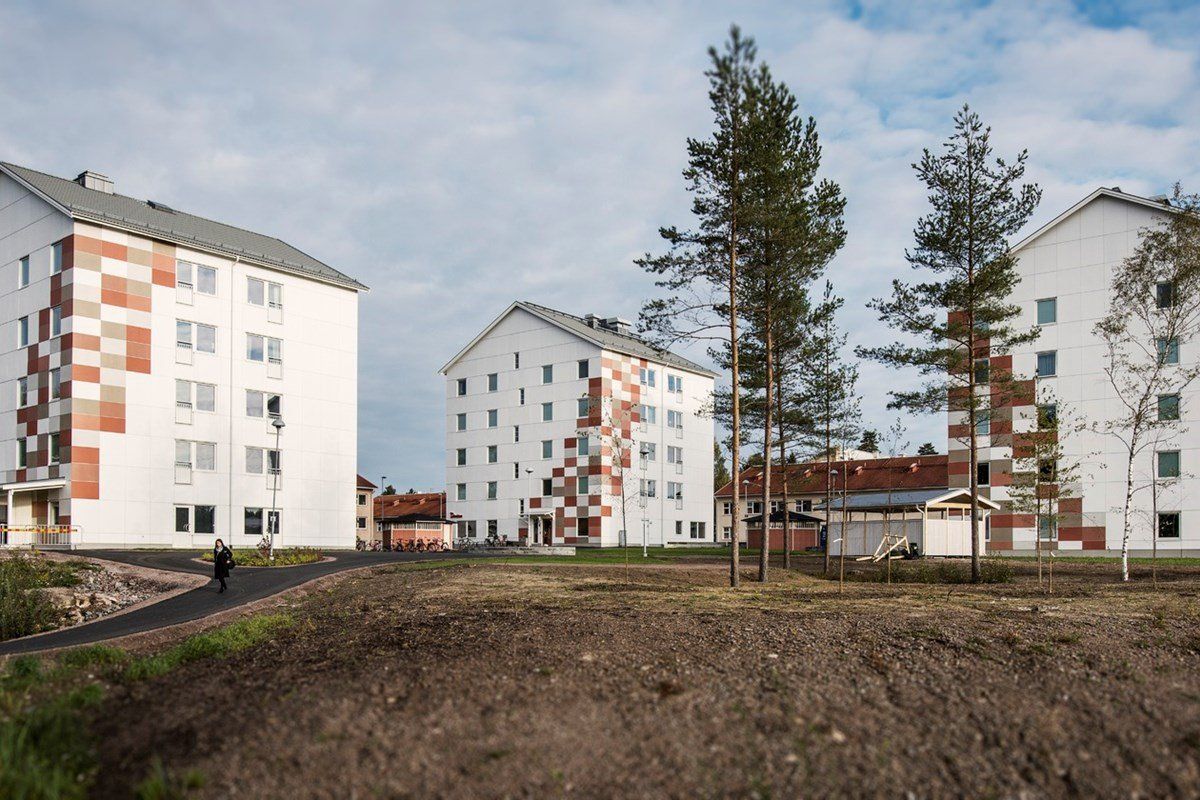 White apartment buildings with colored accents on a grassy lot, trees, and a person walking on a path under a cloudy sky.