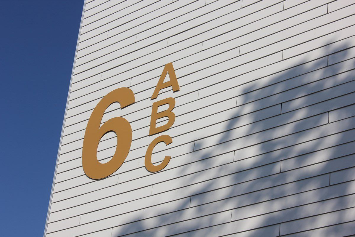 Gold numbers and letters (6, A, B, C) on white brick wall. Shadows of leaves on the wall. Blue sky in the background.