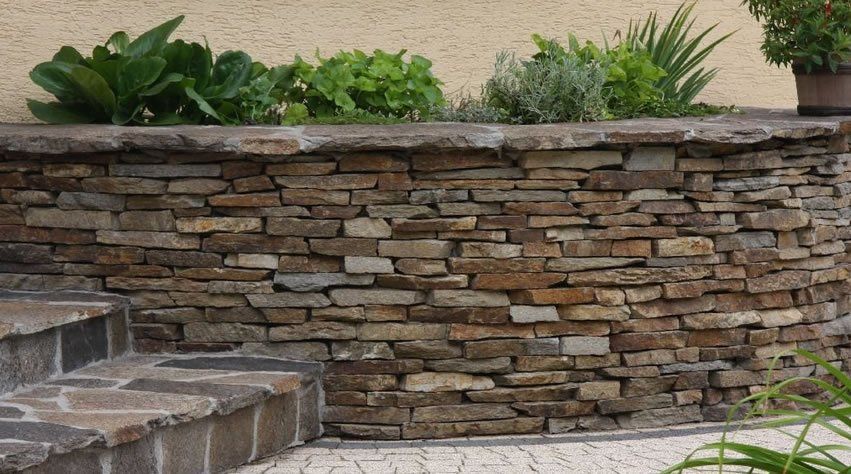 Stone retaining wall with plants growing along the top, built next to stone steps and a patterned path.