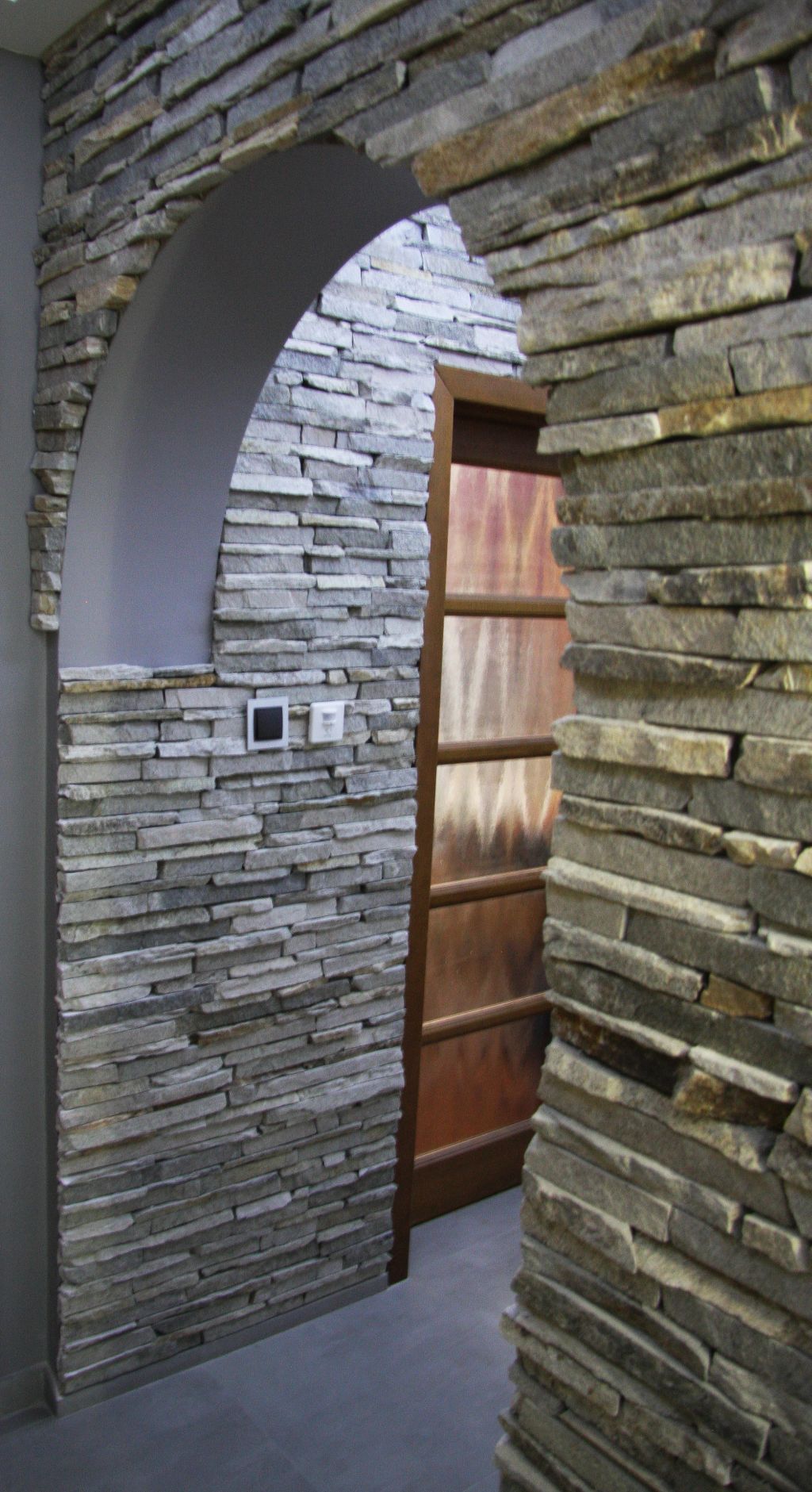 Stone-clad archway leads to a wooden door, grey walls, with electrical outlet visible.