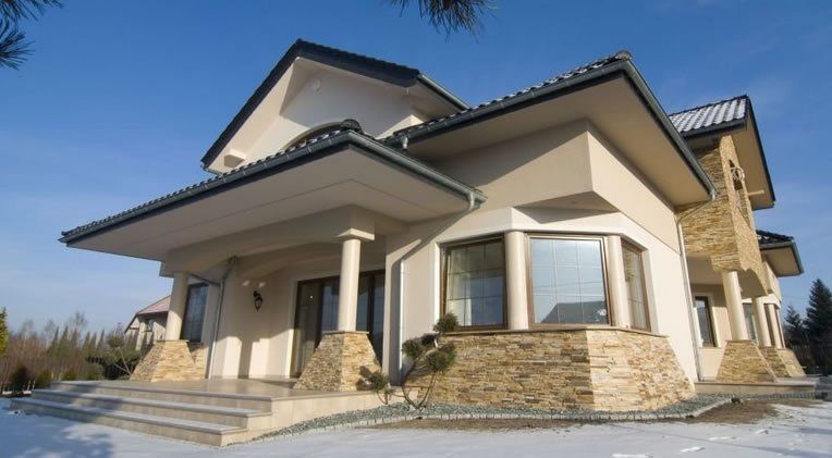 Beige and stone two-story house with columns, snow on the ground, and a clear blue sky.