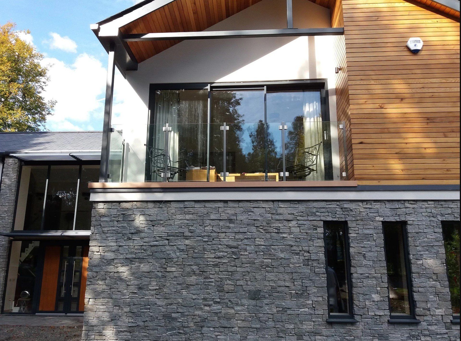 Modern house exterior with stone and wood siding, glass balcony, and large windows.