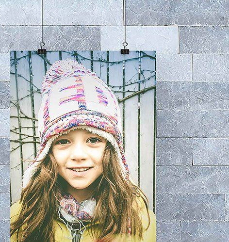 Smiling girl in a winter hat, scarf, and yellow coat, against a blurry fence background.