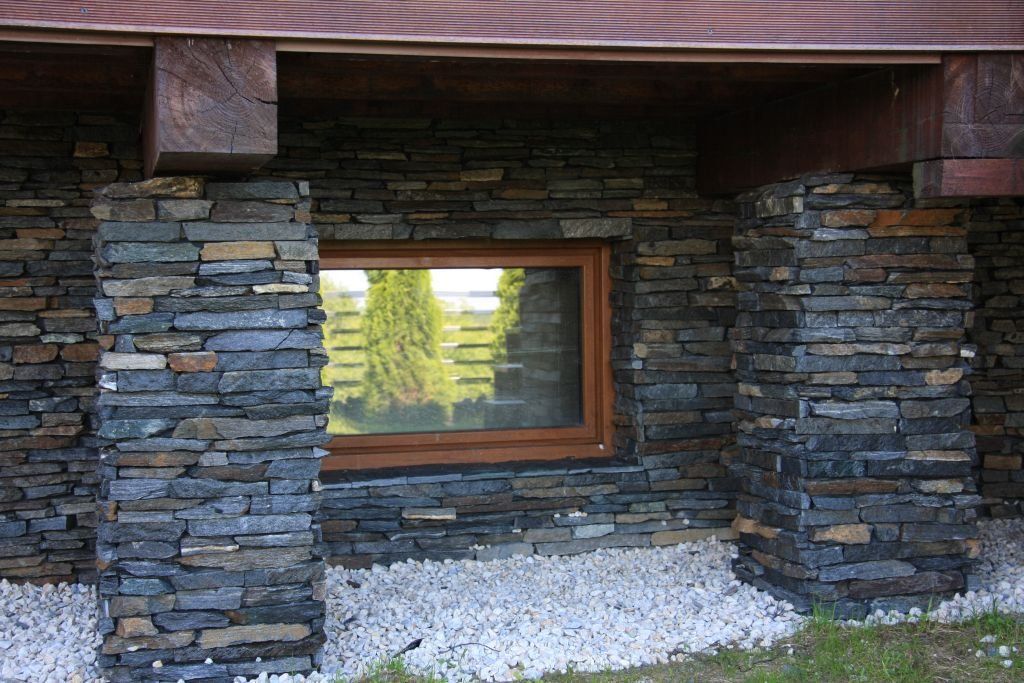 Stone facade with window and wooden beams, surrounded by white gravel and green grass.