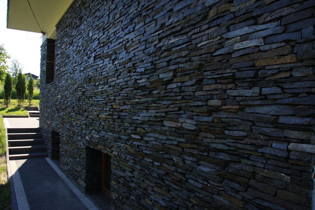 Dark stone facade of a building, with a few small windows and a set of stairs leading to a side entrance.