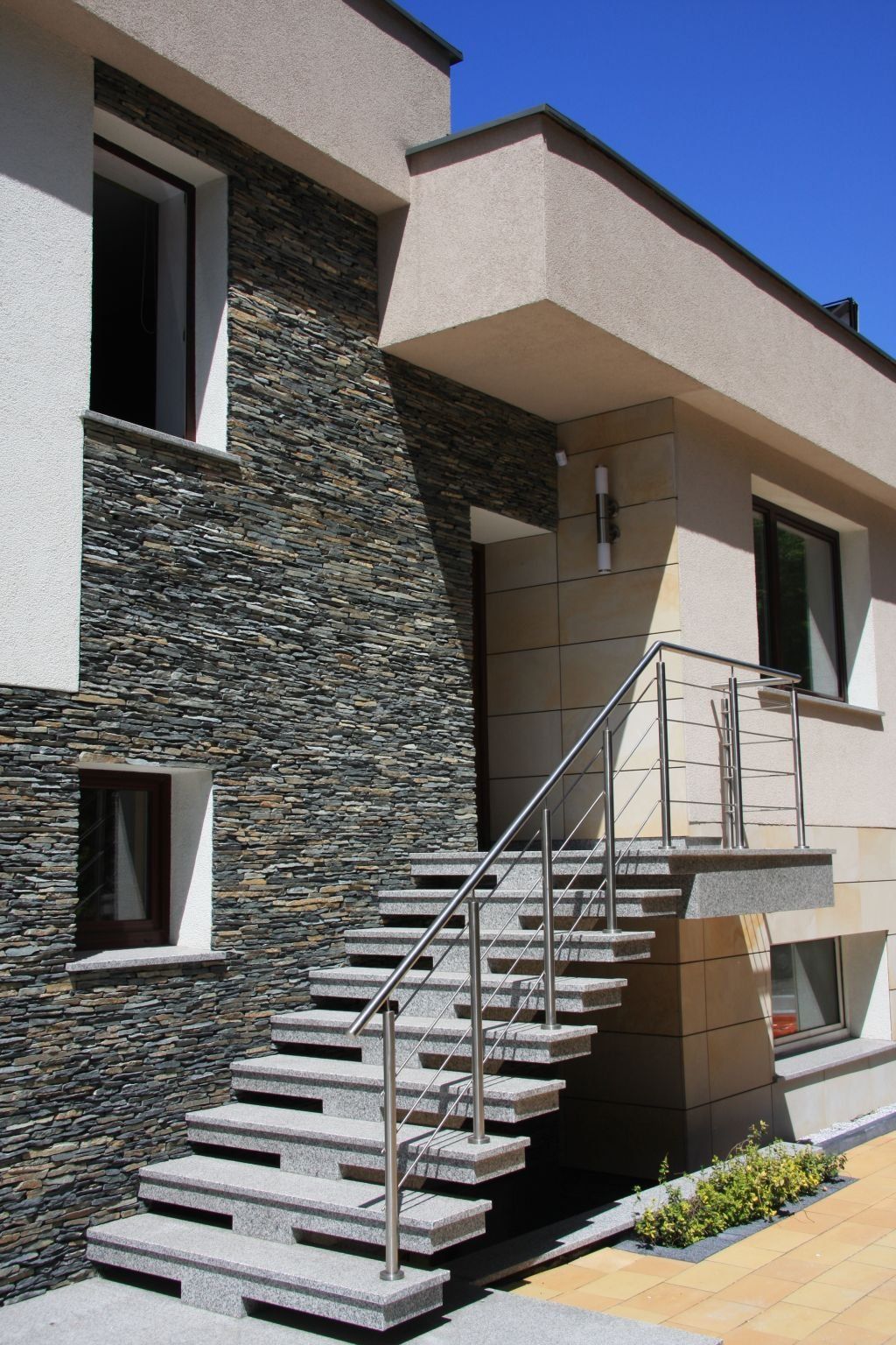Exterior of a modern building with stone wall, stairs, and metal railing. Bright sunlight.