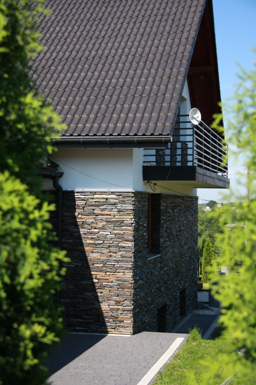 Stone-clad house with dark tile roof and small balcony, partially obscured by green trees.