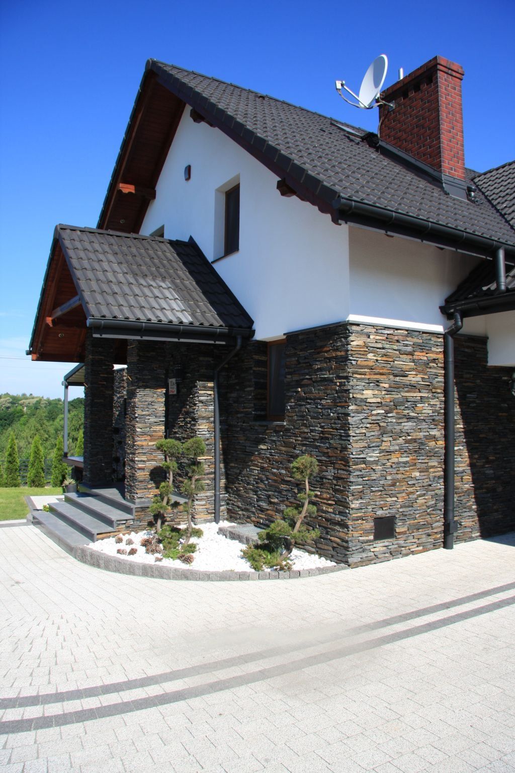 Two-story house with black roof, stone facade, and paved driveway on a sunny day.