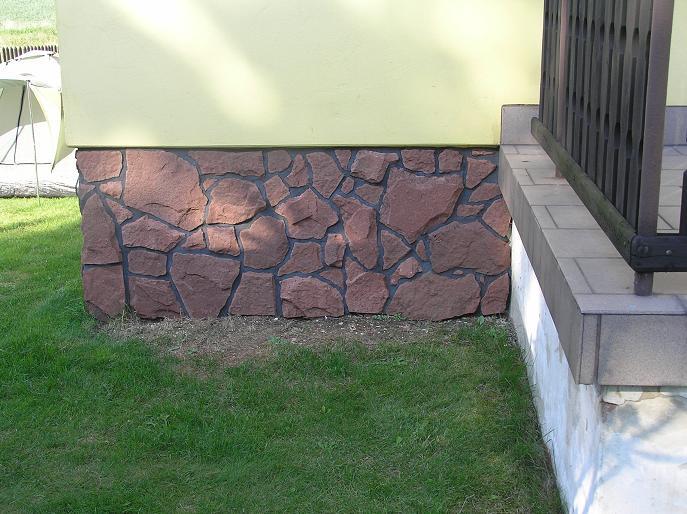 Stone facade on a house foundation; brown stone with black grout against a green lawn and light yellow wall.