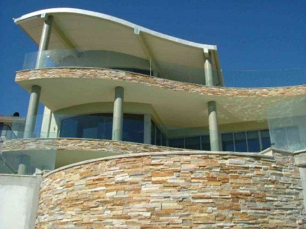 Modern house with stone facade, curved roof, glass railings, and blue sky.