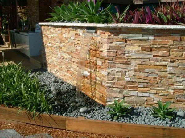 Water feature with stone facade, overflowing into a bed of dark grey rocks, plants in wooden border.