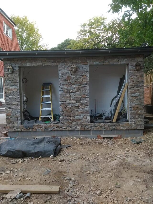 Stone-faced, two-bay structure under construction. Yellow ladder inside left bay, building materials visible. Gray, overcast day.