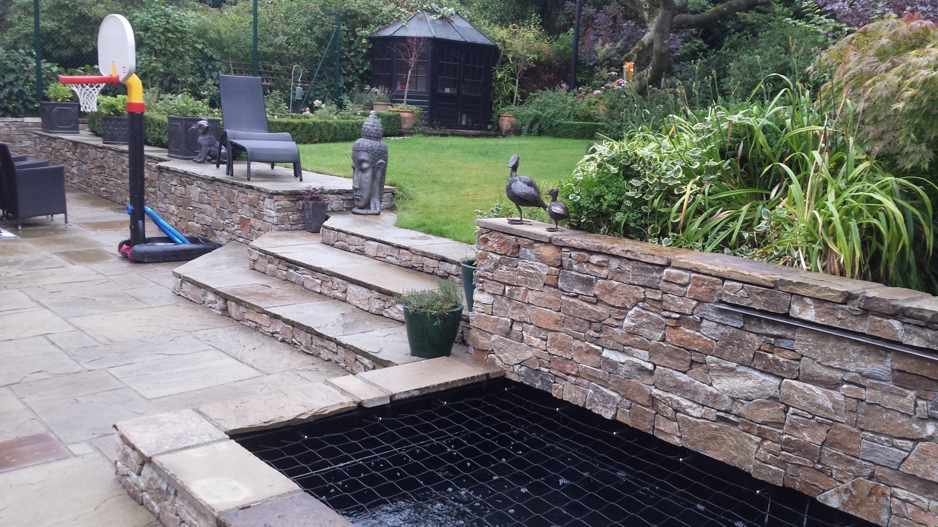 Stone-walled backyard with a fish pond, steps, and a grassy lawn; a basketball hoop is also present.
