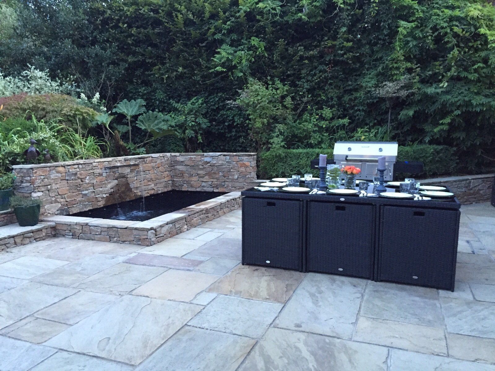 Outdoor dining area with a grill and pond. Black table and chairs on a stone patio surrounded by greenery.