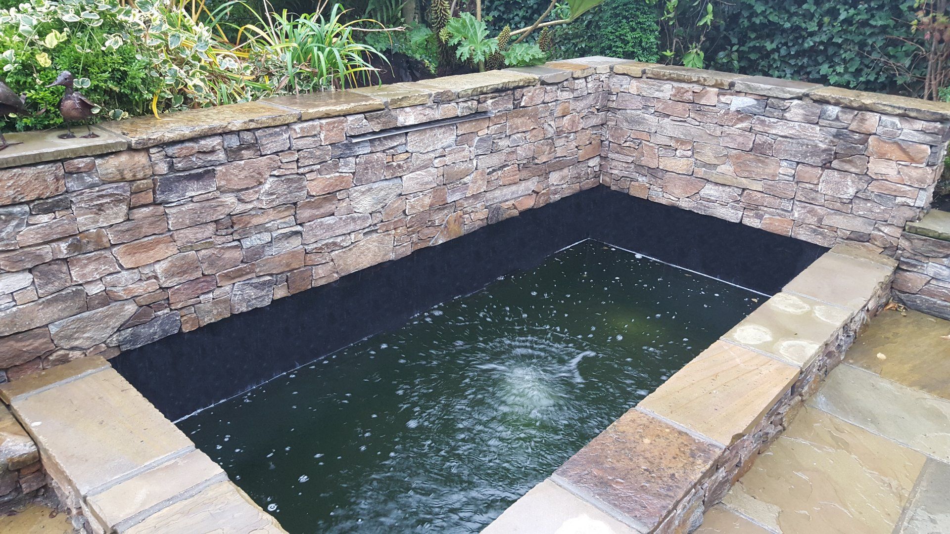 Stone-walled rectangular pond with dark liner and water fountain; set in patio.