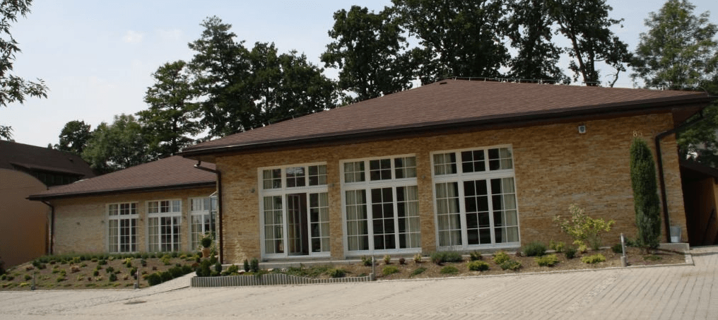 A light-colored brick building with multiple large windows and a brown roof is shown on a sunny day.