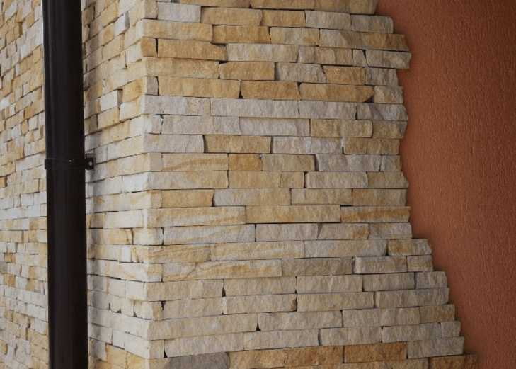 Corner of a building with light tan stone veneer and a dark brown downspout; adjacent wall is reddish-brown stucco.