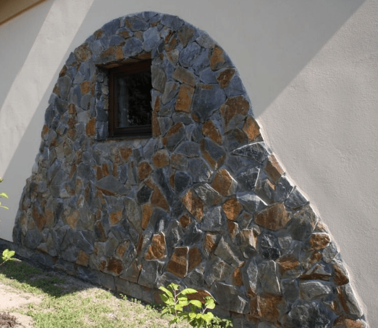 Stone facade arch with small window, contrasting with white wall.