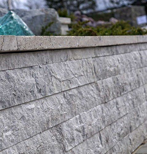 Gray stacked stone retaining wall with a flat cap, partially obscured plants in the background.