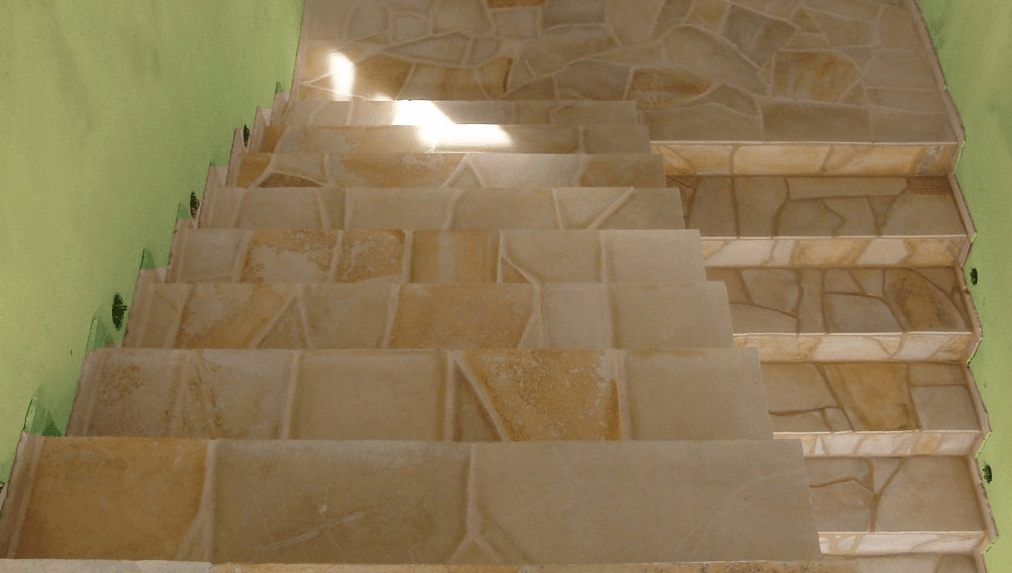 Staircase with light brown stone tiles, descending between green walls; sunlight visible.