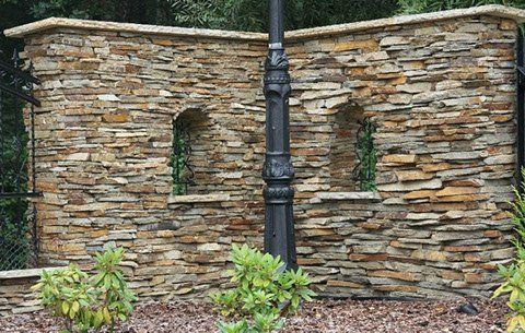 Stone wall with arched niches and a black lamppost. Green plants in front.