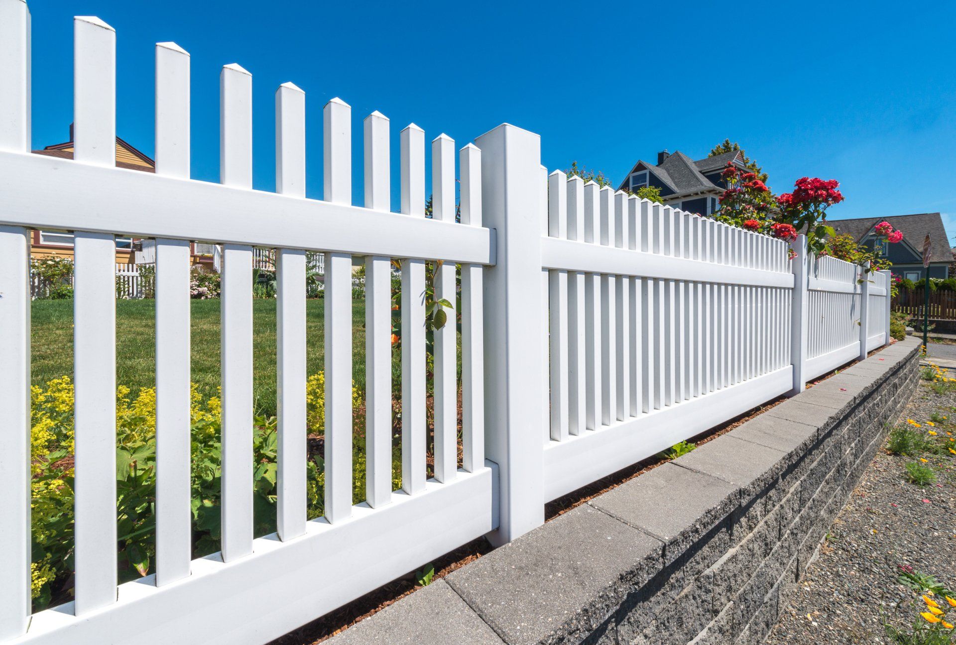 A white picket fence is surrounded by flowers and a stone wall.