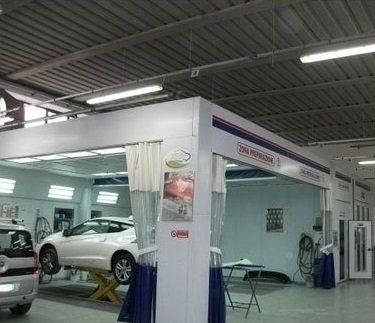 Officina meccanica con un'auto argentata sul ponte sollevatore e postazioni di servizio illuminate da luci fluorescenti.